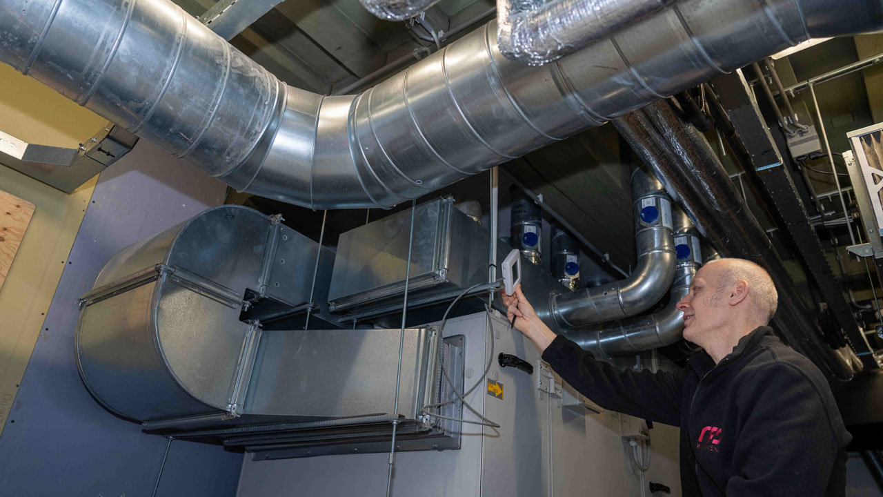 Technicus inspecteert metalen luchtkanalen in een industriële ruimte met meetapparaat, onderdeel van HVAC-onderhoud binnen vastgoedbeheer