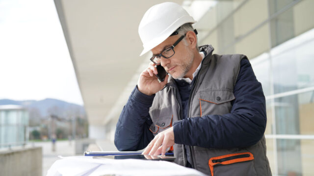 Inspecteur met witte helm bekijkt bouwtekeningen en telefoneert bij moderne gebouwen op de achtergrond