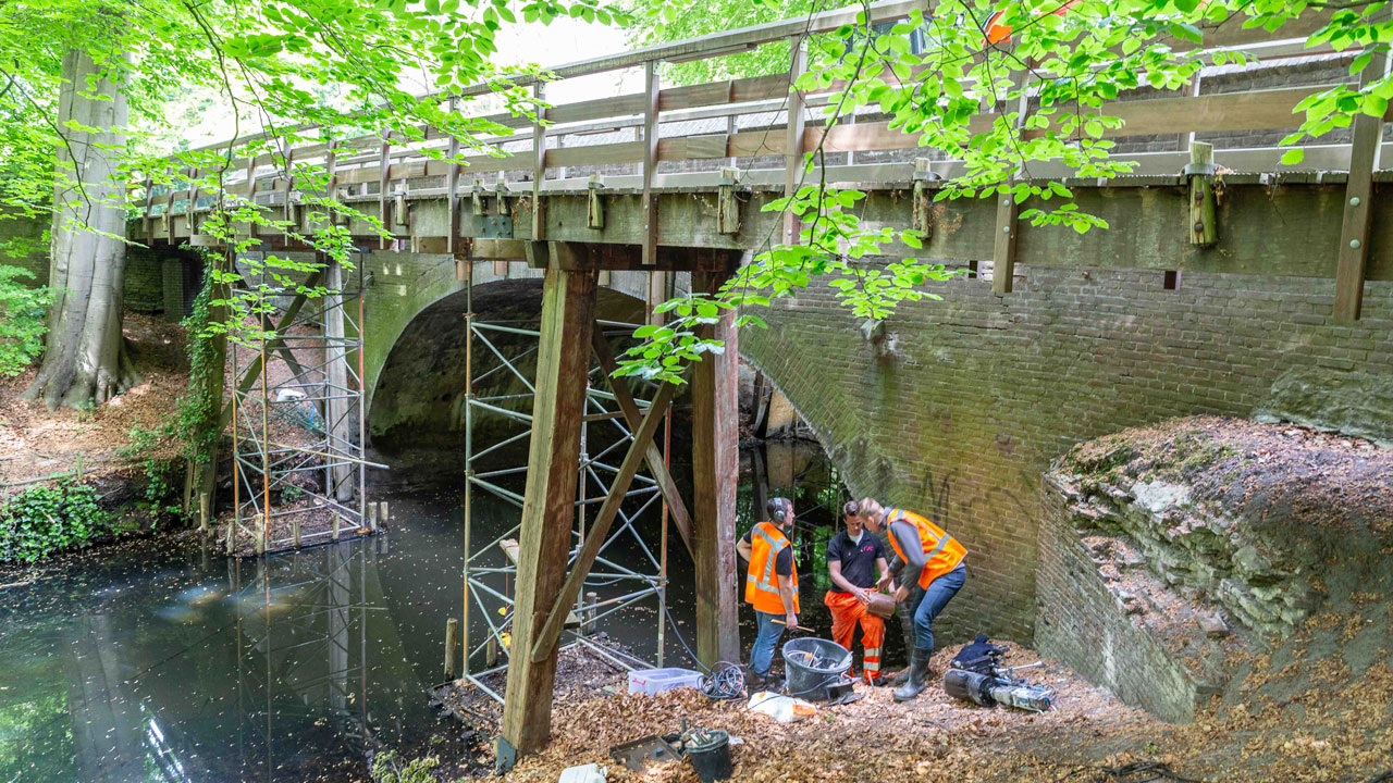 Tetra Tech voert onderhoud uit aan rijksmonumentale boogbrug in Naarden, met steigers en materieel in groene, waterrijke omgeving