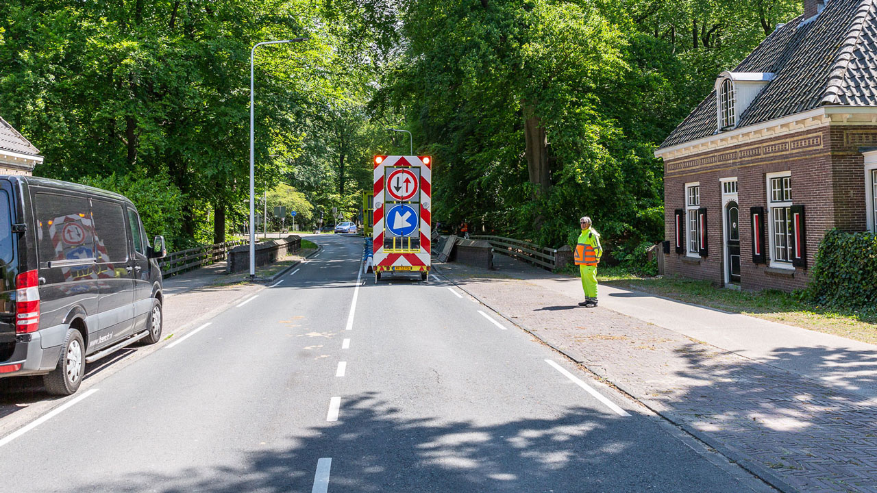 Tetra Tech-voertuig en medewerker bij weginspectie nabij rijksmonumentale boogbrug in Naarden, met waarschuwingsborden en groene omgeving