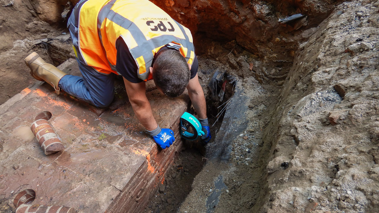 Onderzoek in sleuf bij rijksmonumentale boogbrug in Naarden, met zicht op bakstenen en leidingen, uitgevoerd door specialist in veiligheidsvest