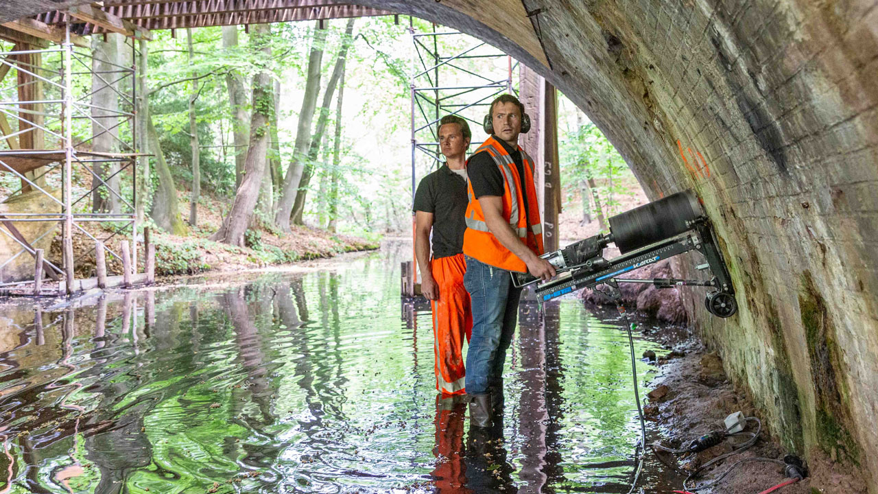 Medewerkers werken onder boogbrug in Naarden, met apparatuur en steiger in ondiep water, omgeven door groen