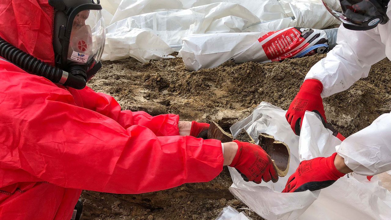 Medewerkers in beschermende pakken verpakken asbesthoudend materiaal uit gresriool in plastic, op locatie in risicoklasse 1