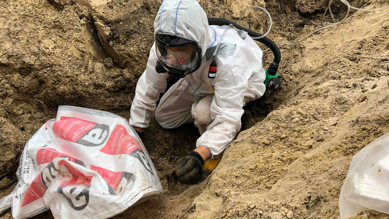 Medewerker in beschermend pak verpakt asbesthoudend materiaal uit gresriool in plastic zak op saneringslocatie risicoklasse 1