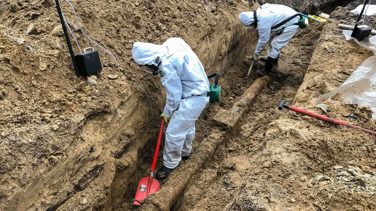 Medewerkers saneren asbesthoudende kit in gresriool, met beschermende kleding en gereedschap in gegraven sleuf