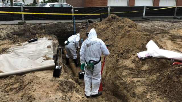 Twee inspecteurs in beschermende pakken voeren een asbestinspectie uit in een bouwput, met woningen en waarschuwingslint op de achtergrond