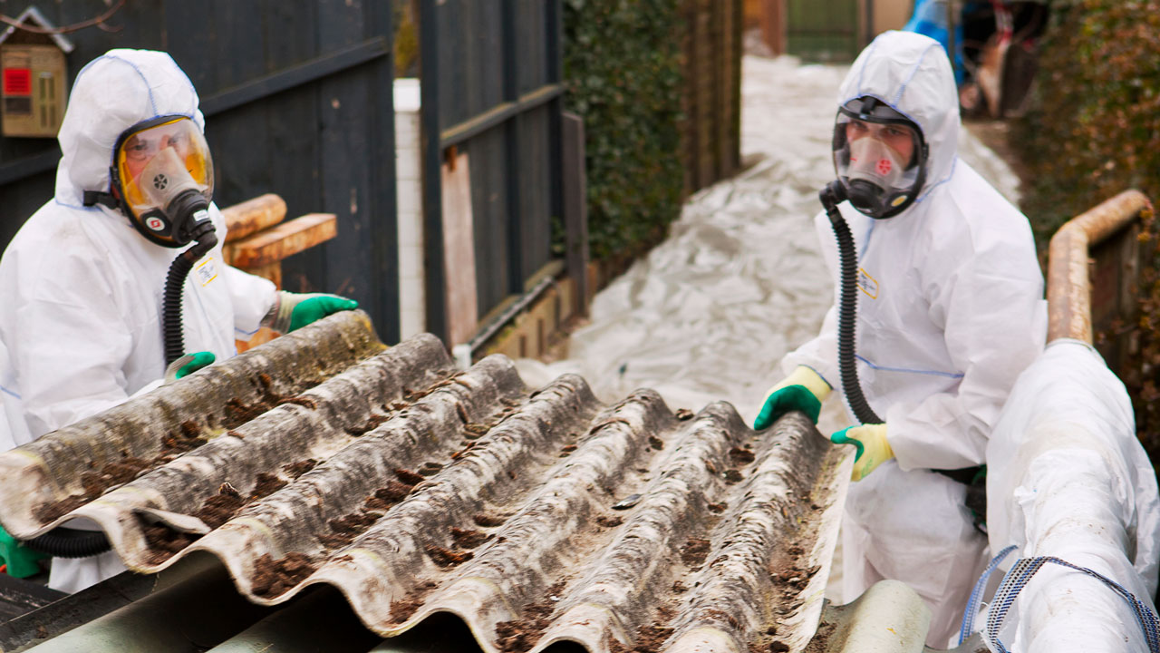 Twee professionals in beschermende pakken verwijderen golfplaten met asbest in woonwijk en plaatsen deze veilig in een met folie beklede container