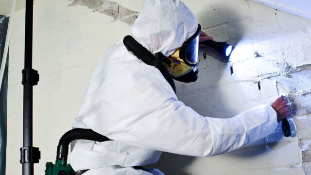 Inspecteur in beschermend pak en masker onderzoekt beschadigde muur met zaklamp tijdens asbestinventarisatie in een mogelijk risicovolle omgeving