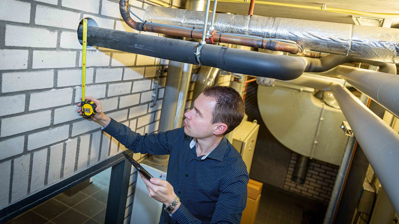 Inspecteur meet een muur in een technische ruimte met leidingen en isolatie, terwijl hij gegevens noteert op een tablet of clipboard