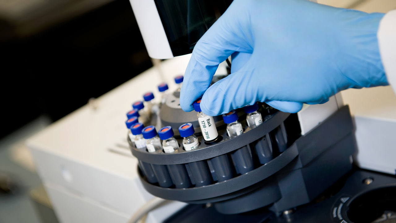 Gehandschoende hand plaatst monsterflesje in autosampler voor chemische analyse in het laboratorium