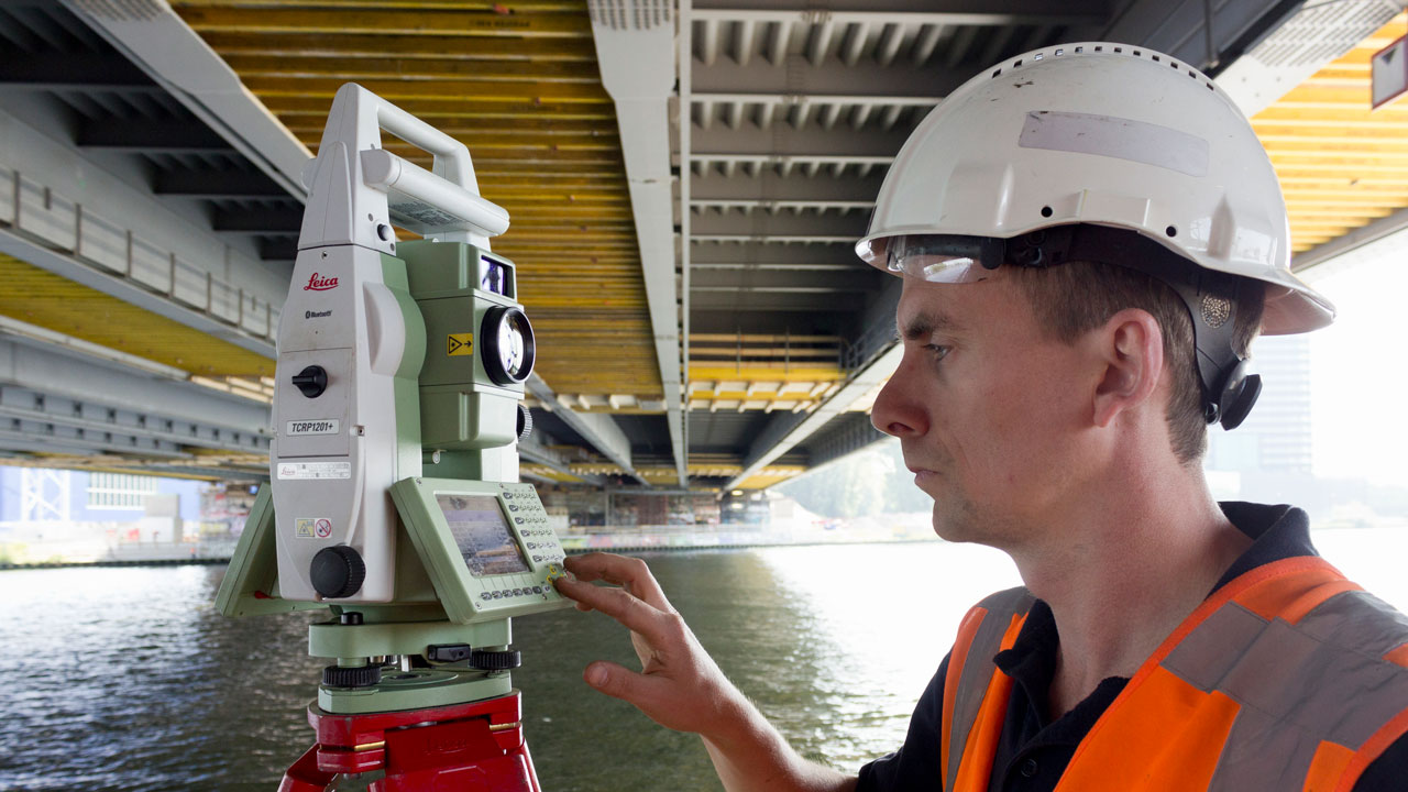 Landmeter met Leica total station voert meting uit onder brug, gericht op monitoring van constructieve vervormingen