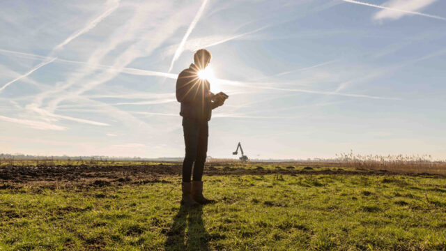 Silhouet van medewerker op grasveld met opkomende zon en graafmachine – duurzame gebiedsinrichting in uitvoering