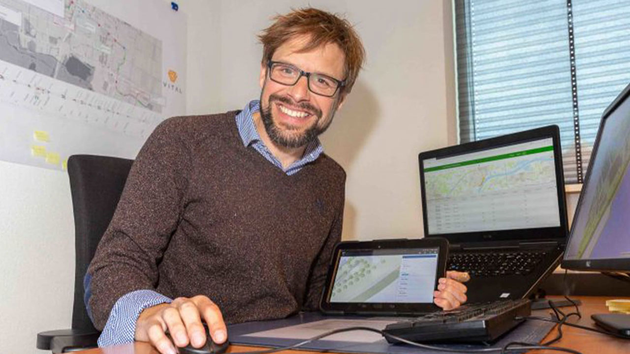 Medewerker analyseert geo-informatie op tablet en meerdere schermen in kantoor met kaart en notities op de muur