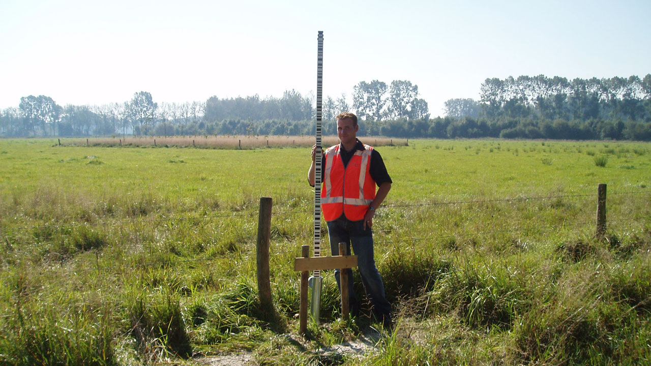 Landmeter met meetstok in grasveld, uitgevoerd met oranje hesje en omringd door meetapparatuur voor hoogtemetingen in het veld