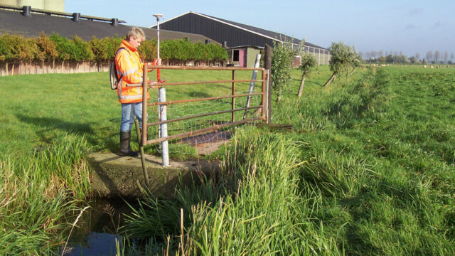 Landmeter op brug met meetapparatuur boven watergang in veld, met landbouwgebouw en bomen op de achtergrond