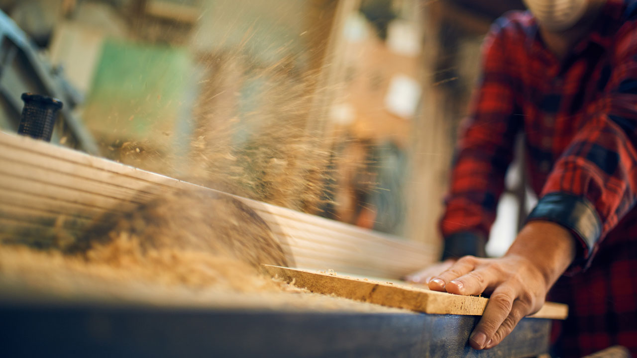 Werknemer zaagt hout in werkplaats met beschermingsmiddelen, waarbij zichtbaar houtstof vrijkomt