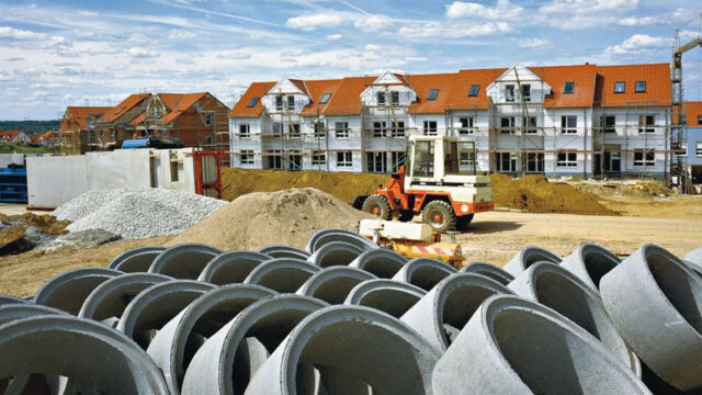 Stedelijke bouwplaats met betonnen rioolbuizen en nieuwbouwwoningen. Wij werken aan infrastructuur en inrichting van woonwijken