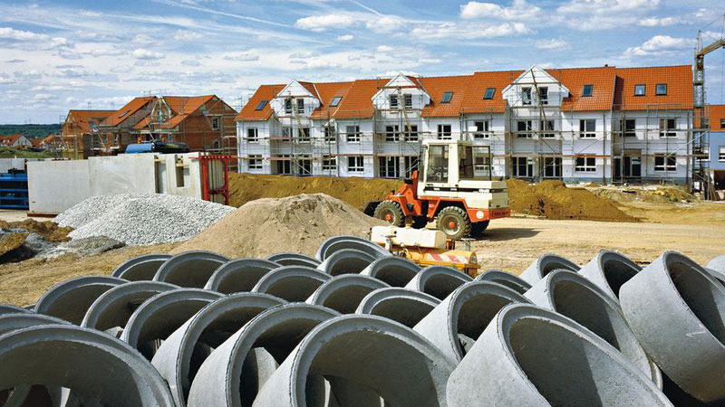 Stedelijke bouwplaats met betonnen rioolbuizen en nieuwbouwwoningen. Wij werken aan infrastructuur en inrichting van woonwijken