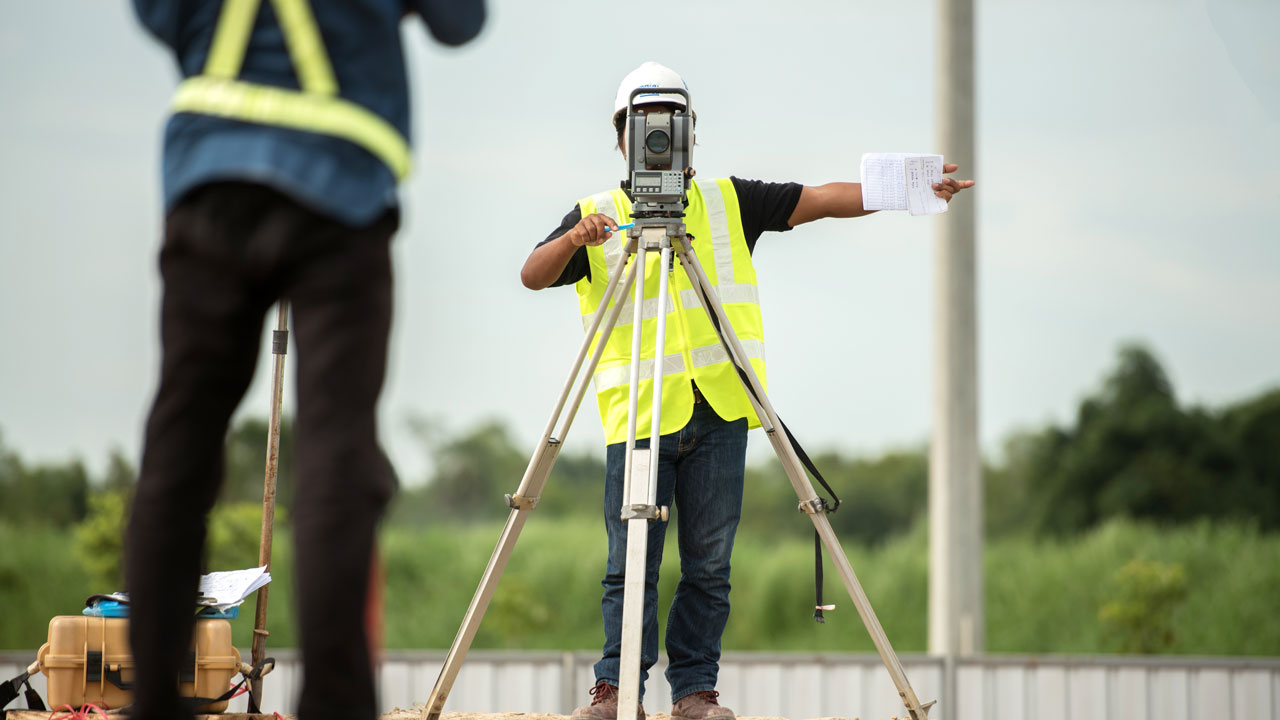 Twee bouwmedewerkers voeren kadastrale meting uit met theodoliet op statief op werklocatie, met helmen en veiligheidsvesten