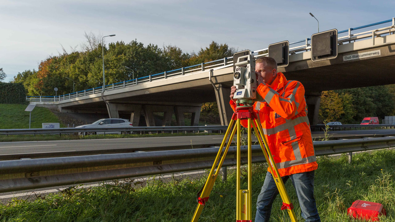 Landmeter in oranje hesje gebruikt meetinstrument op statief langs een weg; op de achtergrond een viaduct, verkeer en bomen