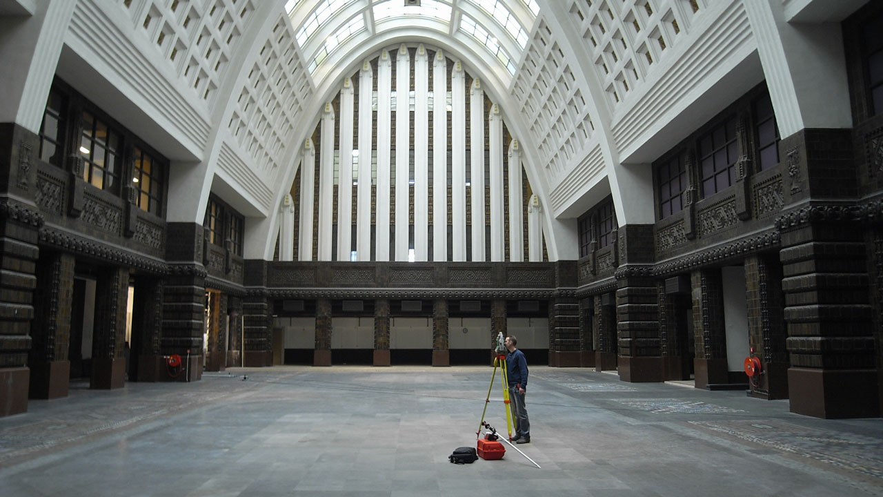 Landmeter voert maatvoering uit in grote hal met gewelfd plafond, natuurlicht en rijke architectonische details