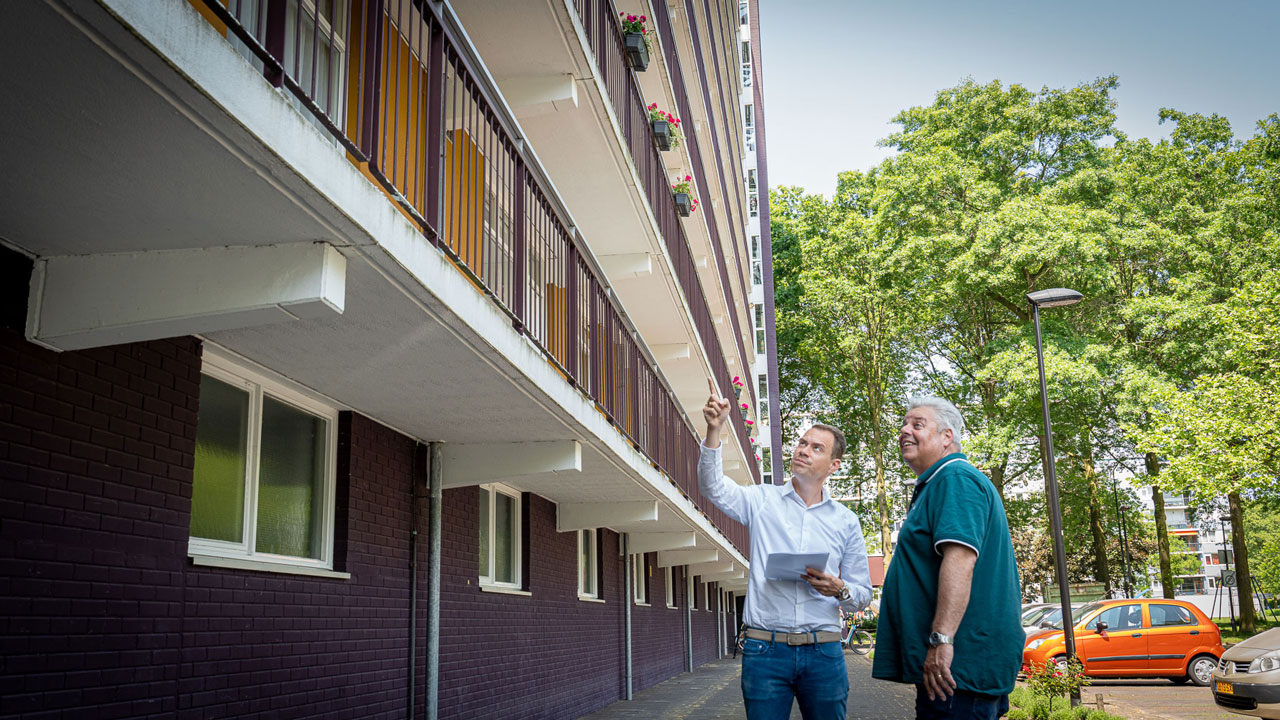 Twee personen inspecteren een woongebouw; één wijst omhoog, de ander houdt een clipboard vast. Op de achtergrond balkons, bomen en geparkeerde auto's