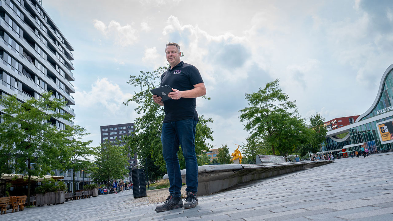 Persoon met tablet in stedelijke omgeving bij het Atlas Theater; op de achtergrond moderne gebouwen, bomen en een deels bewolkte lucht