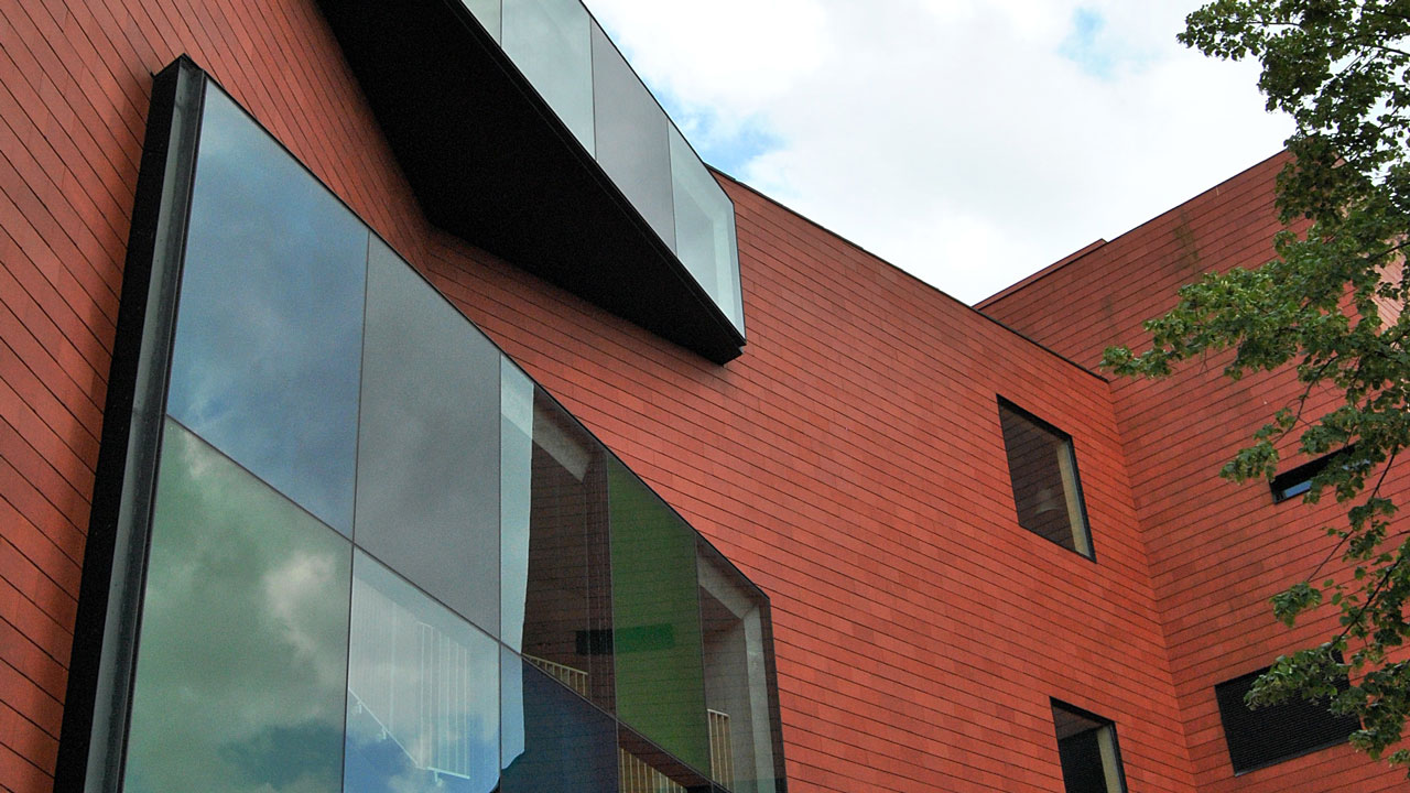 Modern gebouw met rode bakstenen en grote, uitstekende glazen ramen; deels bewolkte lucht en een boom zichtbaar aan de rechterzijde
