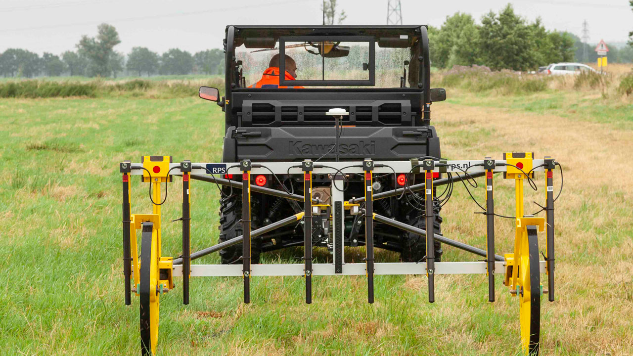 Voertuig met gele sensoren scant grasveld op niet-gesprongen explosieven, met bomen en hoogspanningslijnen op achtergrond