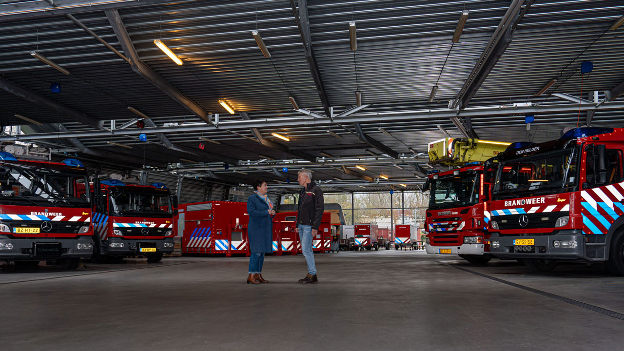 Henny Post en Andre Bult in gesprek tussen brandweerwagens in kazerne Den Helder tijdens brandveiligheidsinspectie.