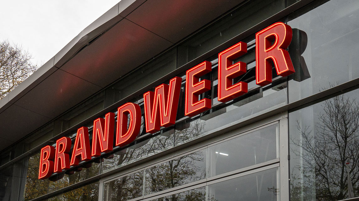 Exterieur brandweerkazerne Den Helder met opvallend rood 'BRANDWEER'-bord; Tetra Tech onderzoekt brandveiligheid en certificering