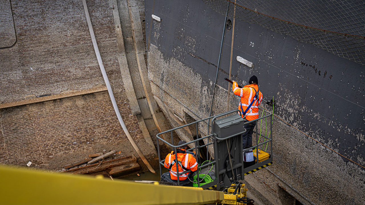 Medewerkers inspecteren droogdokdeur vanaf hoogwerker, onderdeel van maritiem onderhoud in industriële havenomgeving