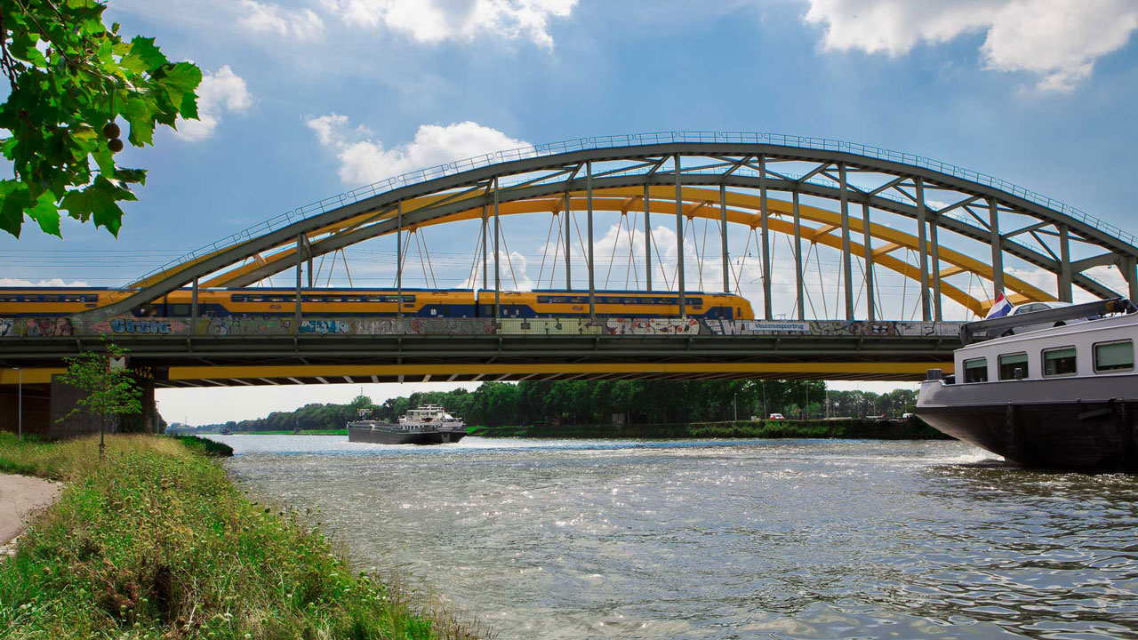 Trein op brug over rivier met boten eronder, illustratie van samenwerking aan digitalisering spoorinfrastructuur door ProRail, Tetra Tech, Geodan en Iv-Infra