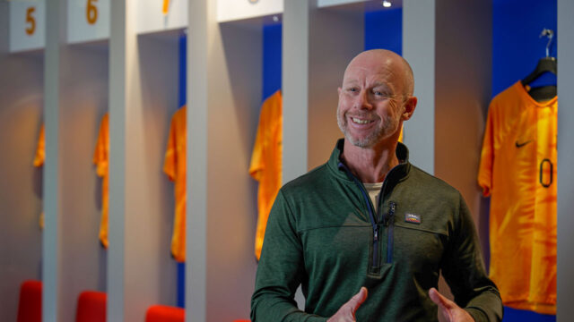 Man spreekt voor sportlockers met oranje shirts, symbool voor teamgeest en balans op de werkvloer binnen organisaties