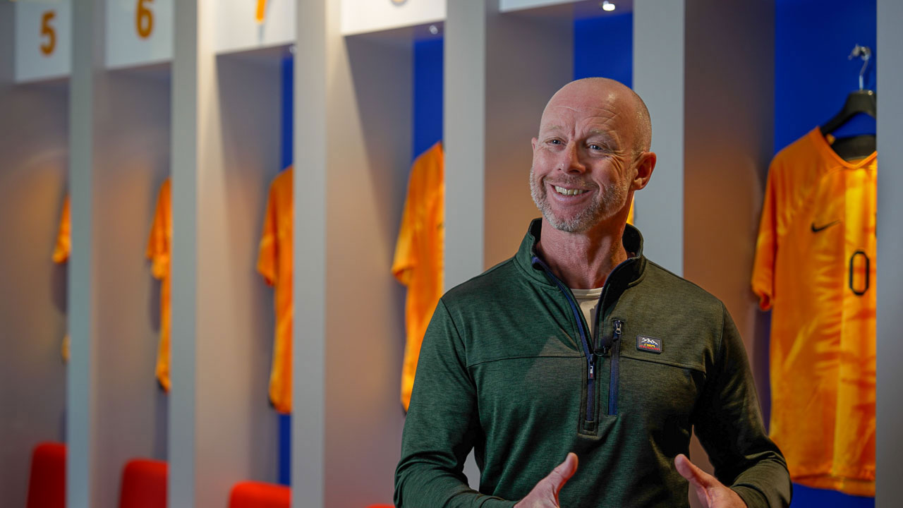 Man spreekt voor sportlockers met oranje shirts, symbool voor teamgeest en balans op de werkvloer binnen organisaties