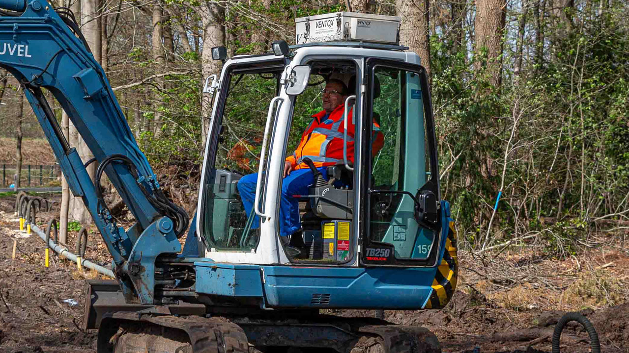 Graafmachine met Tetra Tech-medewerker op modderige bouwplaats N226, aanleg kabels en leidingen in bosrijke omgeving