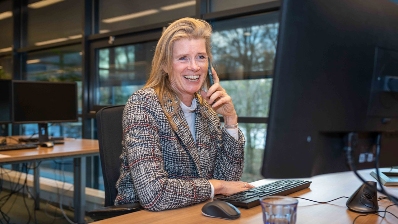 Projectleider Karla van Stekelenburg aan het werk op kantoor, schakelt tussen telefoon en computer bij voorbereiding van bouwkundige inspecties en rapportages
