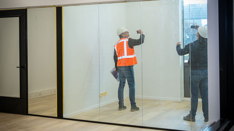 Medewerkers met helm en vest inspecteren glazen wand, illustratie van kwaliteitscontrole bij duurzaam bouwen in Schiedam
