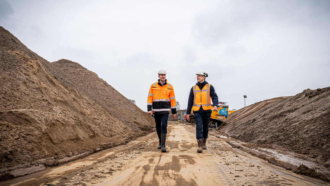 Medewerkers lopen over modderige bouwplaats met grondverzet – onderdeel van project De Dijk van de Toekomst