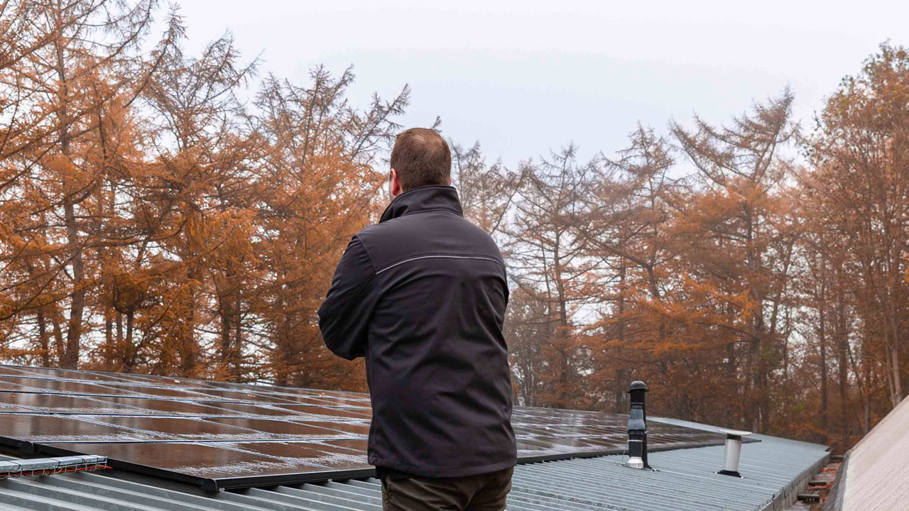 Persoon op dak met zonnepanelen, rug naar camera, herfstbomen op de achtergrond; focus op duurzaam onderhoud en energiebeheer