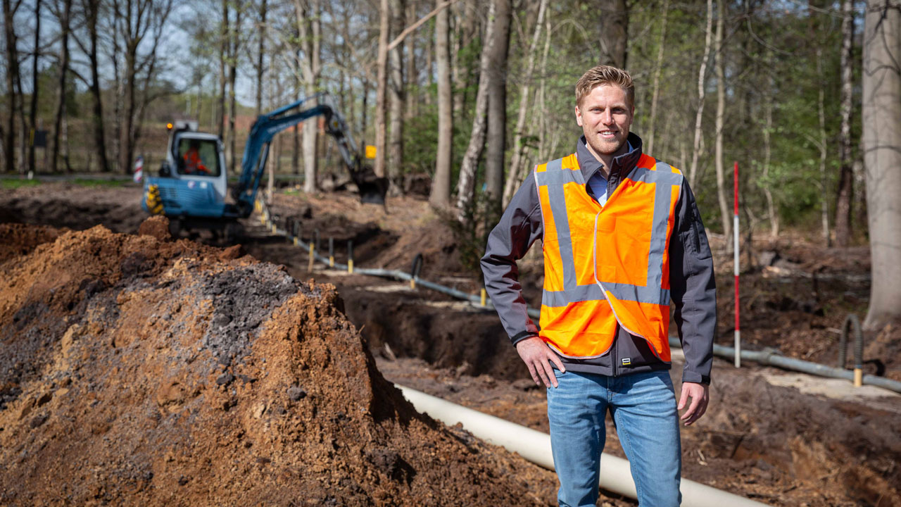 Bouwplaats N226 met Tetra Tech-medewerker en graafmachine bij aanleg kabels en leidingen in bosrijke omgeving