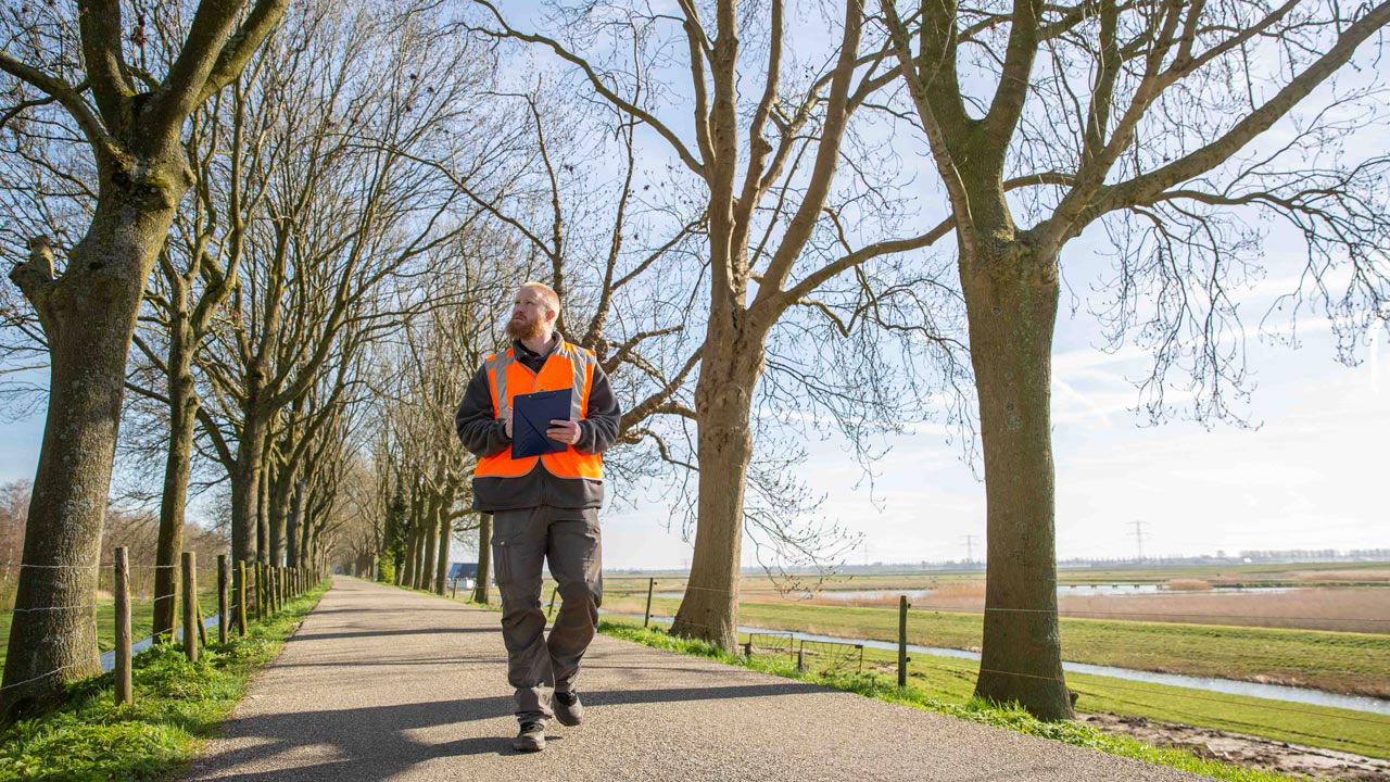 Medewerker in oranje veiligheidsvest met clipboard op pad langs bomenrij en watergang – monitoring en beheer van natuur en infrastructuur door Tetra Tech