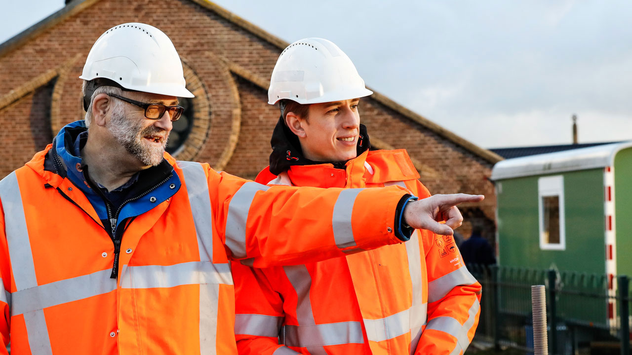 Twee Tetra Tech-medewerkers in veiligheidskleding overleggen op locatie bij een gebouw, met focus op ruimtelijke afstemming
