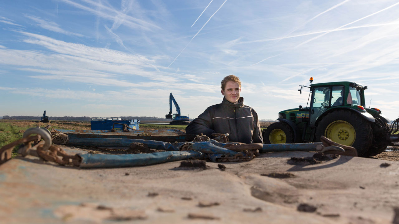 Persoon bij landbouwvoertuig met kettingen en werktuig, illustratie van werkzaamheden in natuurherstelproject Oostelijke Vechtplassen
