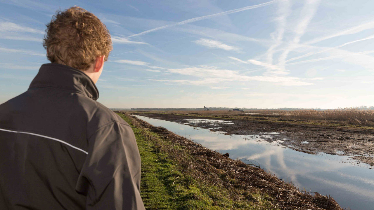 Persoon op graspad langs water in open landschap, illustratie van veldwerk en natuurbeheer in de Oostelijke Vechtplassen
