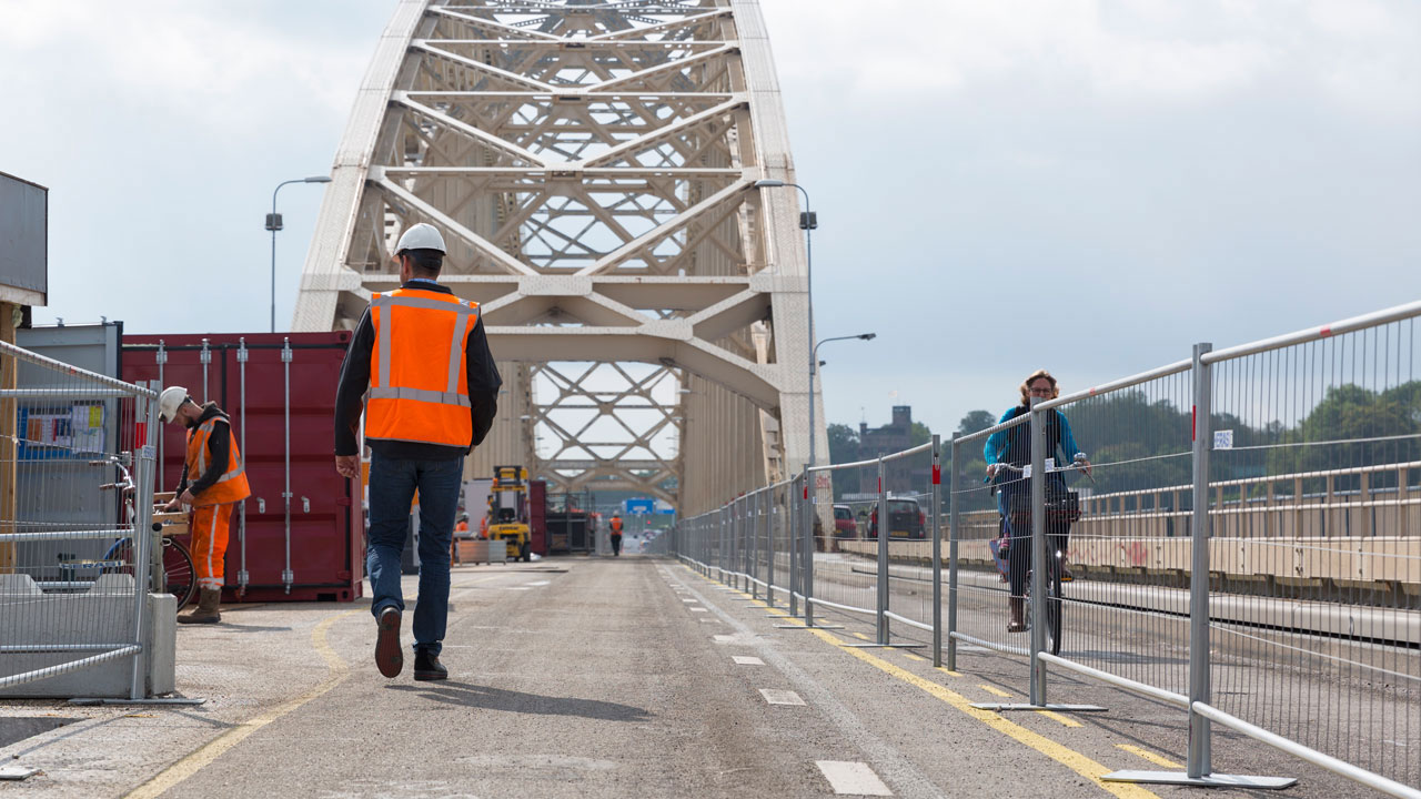 Tetra Tech-medewerker inspecteert brug in onderhoud met bouwvakkers en passerende fietser op de achtergrond