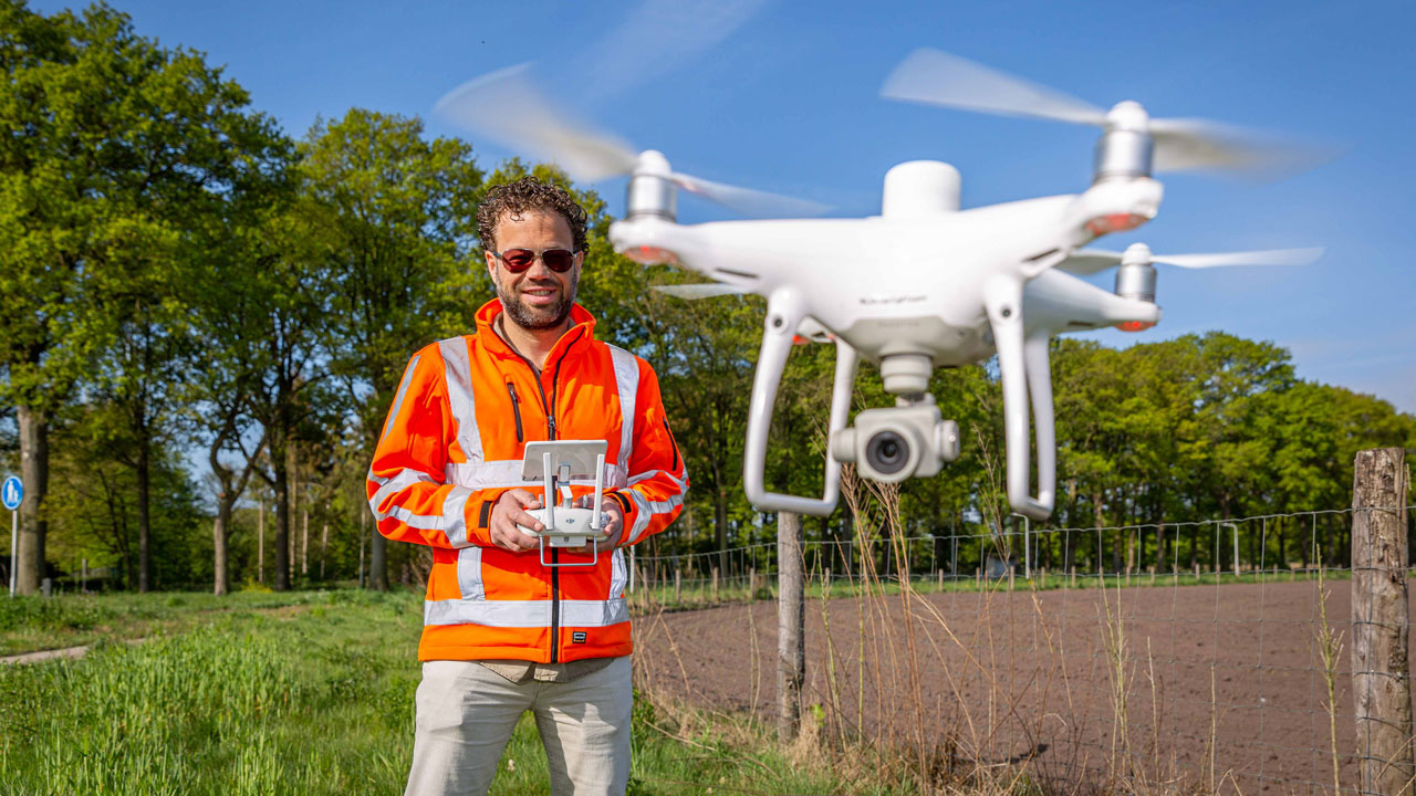 Dronepiloot in oranje veiligheidsjas bestuurt vliegende drone boven open veld – nauwkeurige inspectie van infrastructuur en terrein met drones
