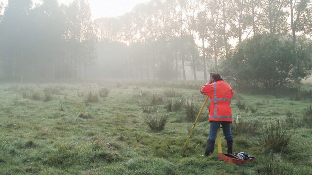 Landmeter met Tetra Tech-jas voert kadastrale meting uit in mistig veld met statief, meetapparatuur en rode gereedschapskist