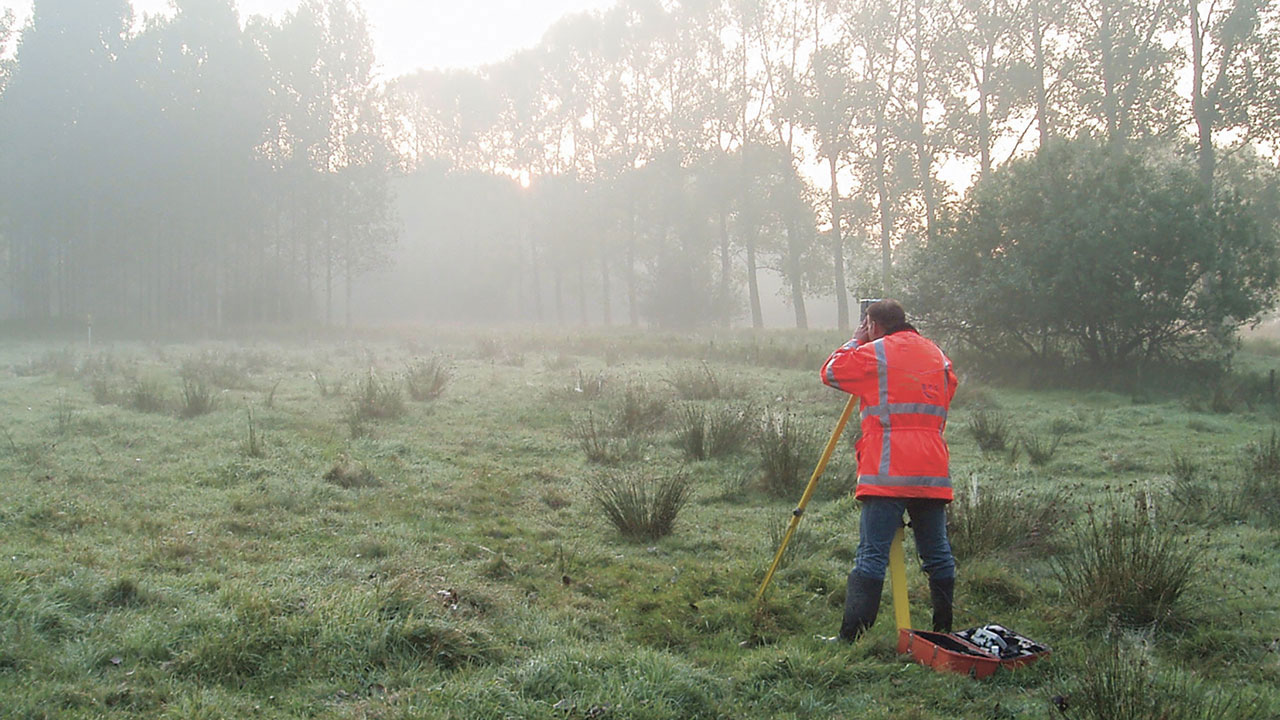 Landmeter met Tetra Tech-jas voert kadastrale meting uit in mistig veld met statief, meetapparatuur en rode gereedschapskist