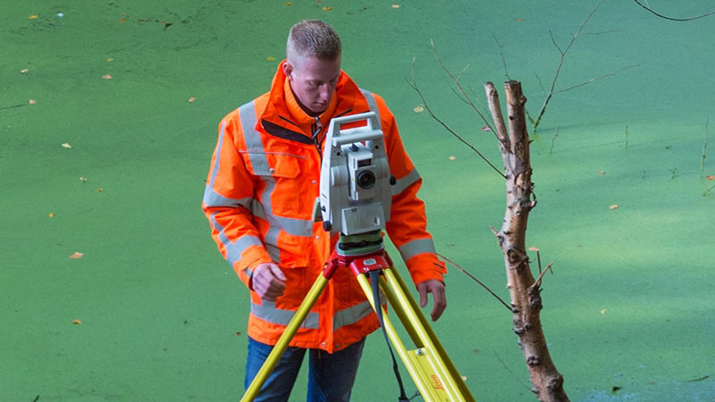 Landmeter met oranje jas werkt met meetinstrument op statief langs waterkant met groene algen.